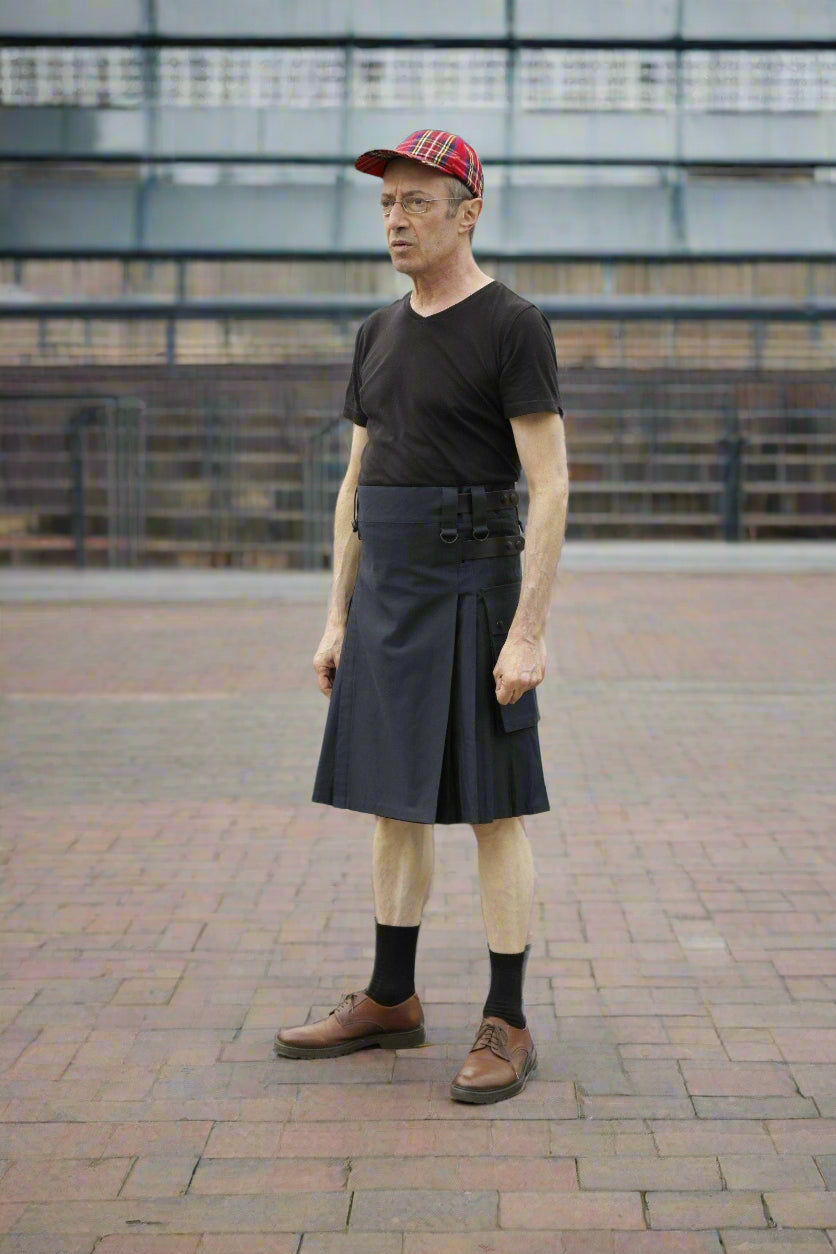 Person wearing a black kilt and cap standing in an outdoor setting with vehicles and a fence in the background.