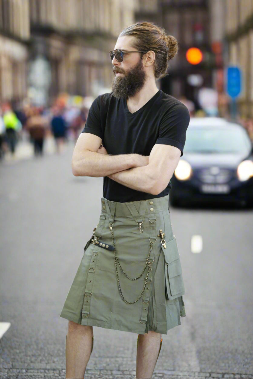 Man wearing a black t-shirt and olive green kilt standing against a brick wall.